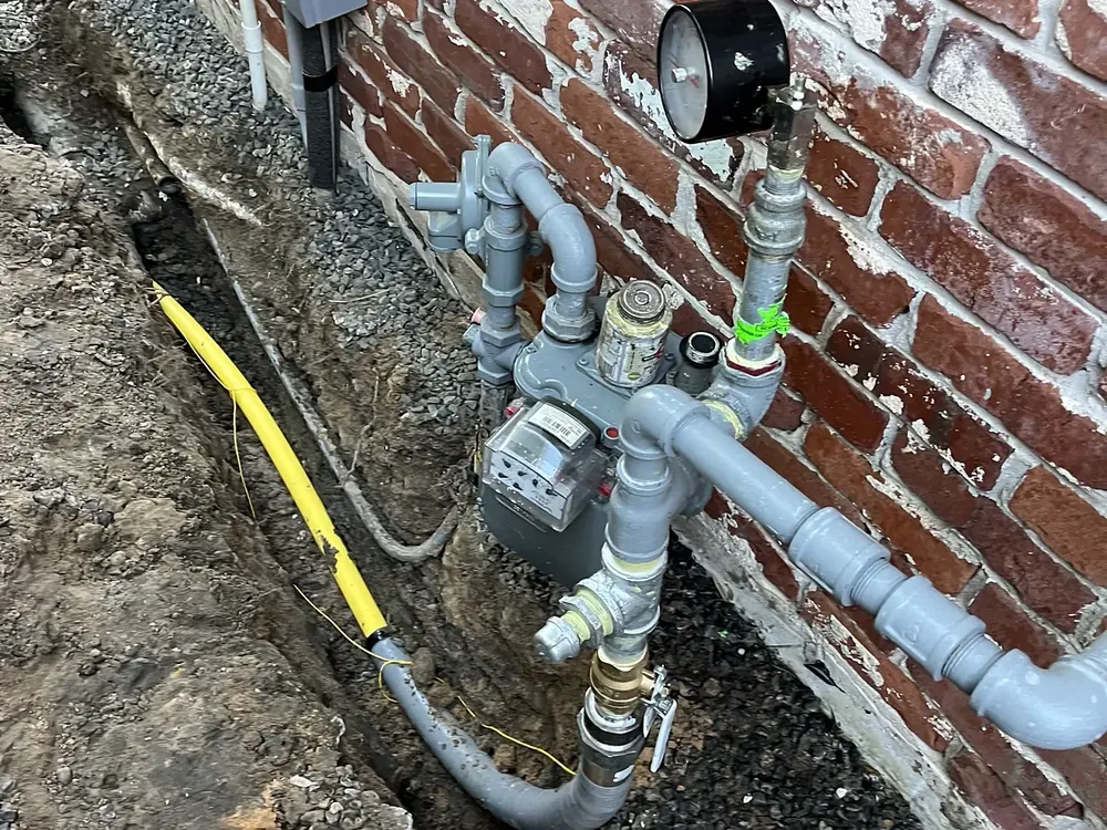 Gas meter set on a brick wall with regulator, pressure gauge, painted steel piping and a yellow gas line in a trench